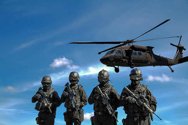 Military police stand in a line as a black helicopter hovers overhead.